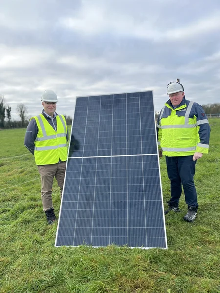 Martin Eager (left) and James Cafferty (right) stand on a green field at Solar Farm Ballycore . A solar panel is positioned between them.