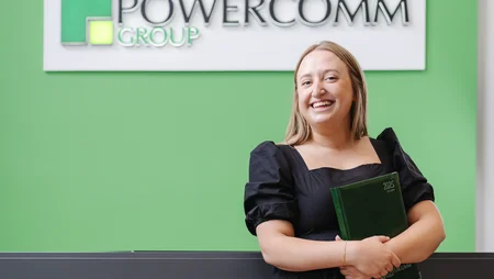 A smiling blonde woman standing in front of reception desk that has a large sign that says Powercomm Group