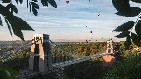 A drone image of the Clifton Suspension Bridge in Bristol by Jarred Kyle, with hot air balloons in the sky and the bridge surrounded by lush greenery